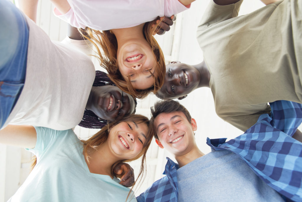 Five smiling adolescents, heads together and facing downwards in a circle with arms locked